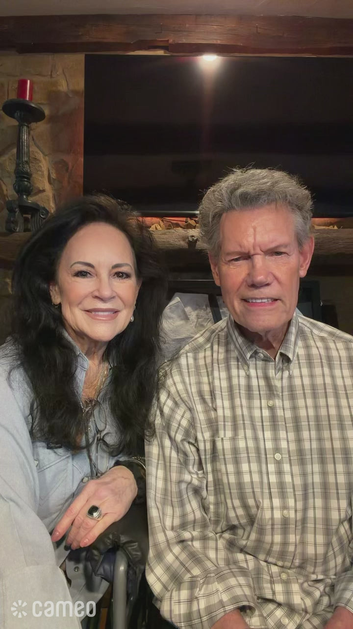 Load video: A vertical Cameo birthday video featuring Randy Travis and a woman sitting side-by-side in a cozy living room.Randy Travis is seated on the right side of the frame. He has gray hair and is wearing a light-colored plaid button-up shirt. The woman is seated beside him on the left. She has long dark hair and is wearing a light gray top.They are sitting in front of a stone fireplace with a wooden mantel and a large dark television above it. The room has a warm, homey appearance. A small “Cameo” watermark appears near the upper-right portion of the video.Throughout the video, Randy and the woman look toward the camera as they speak. They smile, laugh, and turn toward each other at different moments. Randy has several expressive reactions while talking, including smiling broadly and laughing.The video is filmed vertically and is framed fairly closely, showing both people from approximately the waist/chest up.